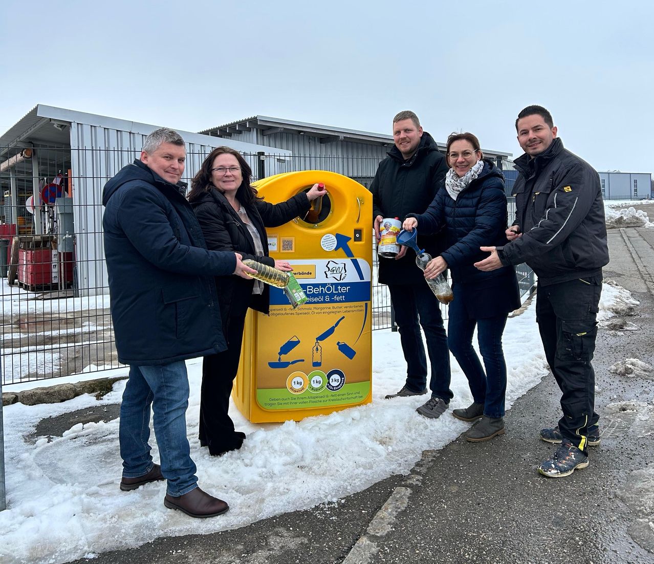 Fünf Personen stehen neben einem gelben Recyclingbehälter, lächeln und halten Flaschen in den Händen. Sie scheinen an einer Recyclingveranstaltung teilzunehmen. Hinter ihnen ist ein Metallzaun und ein Gebäude zu sehen.