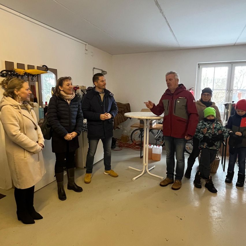 Eine Gruppe von Menschen steht in einem Raum mit einem sprechenden Mann, in der Nähe eines Tisches und eines Fahrrads. Ein Kind mit grüner Mütze hört zu.