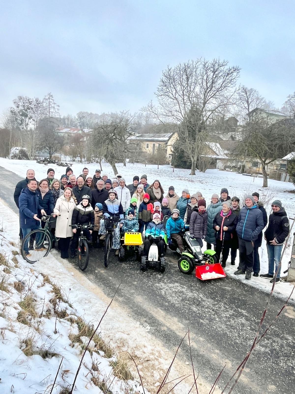 Eine Gruppe von Menschen steht auf einer verschneiten Straße mit Fahrrädern und einem Go-Kart, trägt Winterkleidung und lächelt. Bäume und Häuser sind im Hintergrund zu sehen.