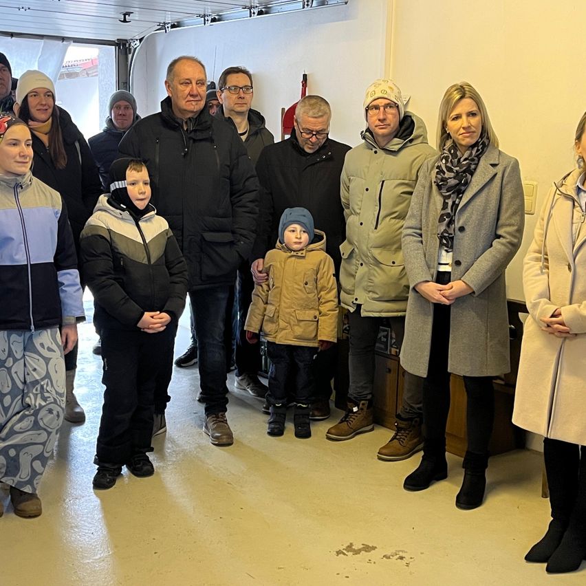 Eine Gruppe von Menschen, einschließlich Kindern, steht in Innenräumen in Winterkleidung. Sie scheinen für ein Foto zu posieren, wobei ein Mann ein Kind hält. Der Raum hat eine weiße Wand und einen Boden.