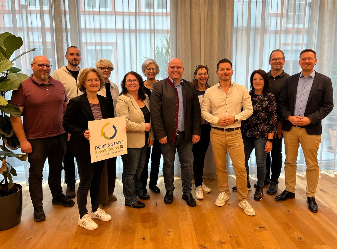 Eine Gruppe von Menschen steht gemeinsam in einem Raum und lächelt für ein Foto. Sie halten ein Schild mit der Aufschrift Dorf & Stadt Erneuerung.