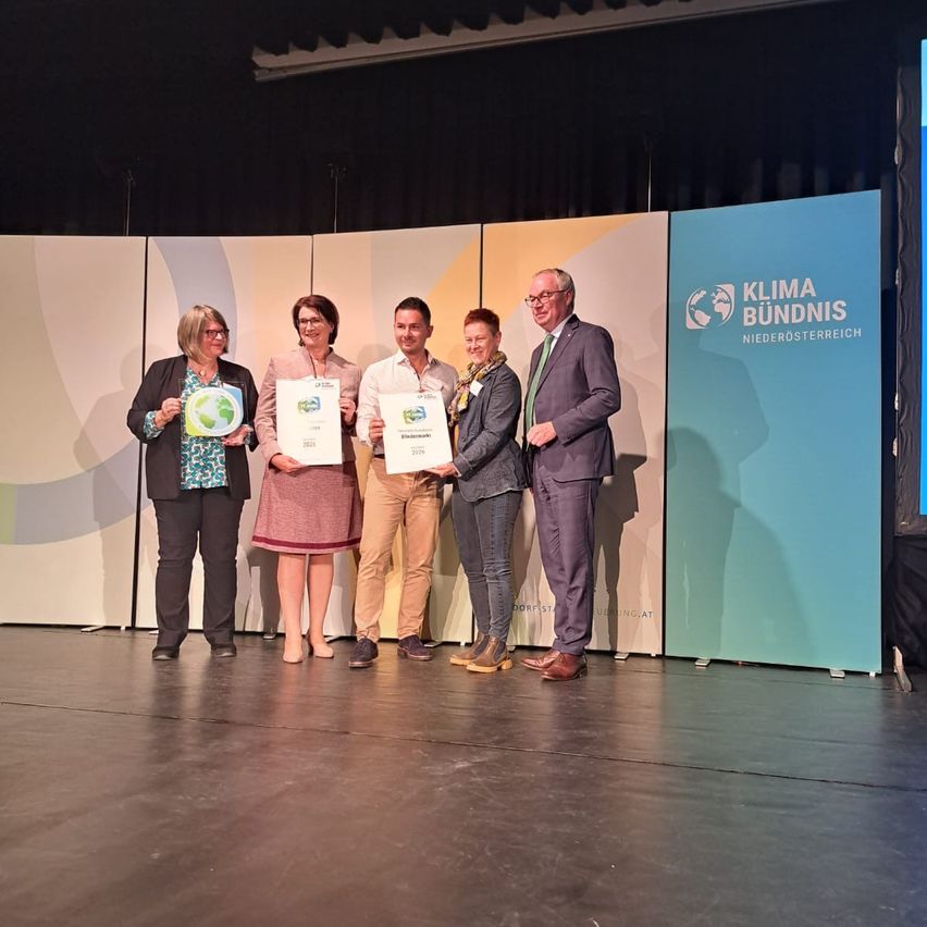 Fünf Personen stehen auf einer Bühne und halten Urkunden in den Händen, vermutlich für eine Preisverleihung. Hinter ihnen sind Banner mit grünen und blauen Designs und Logos angebracht.