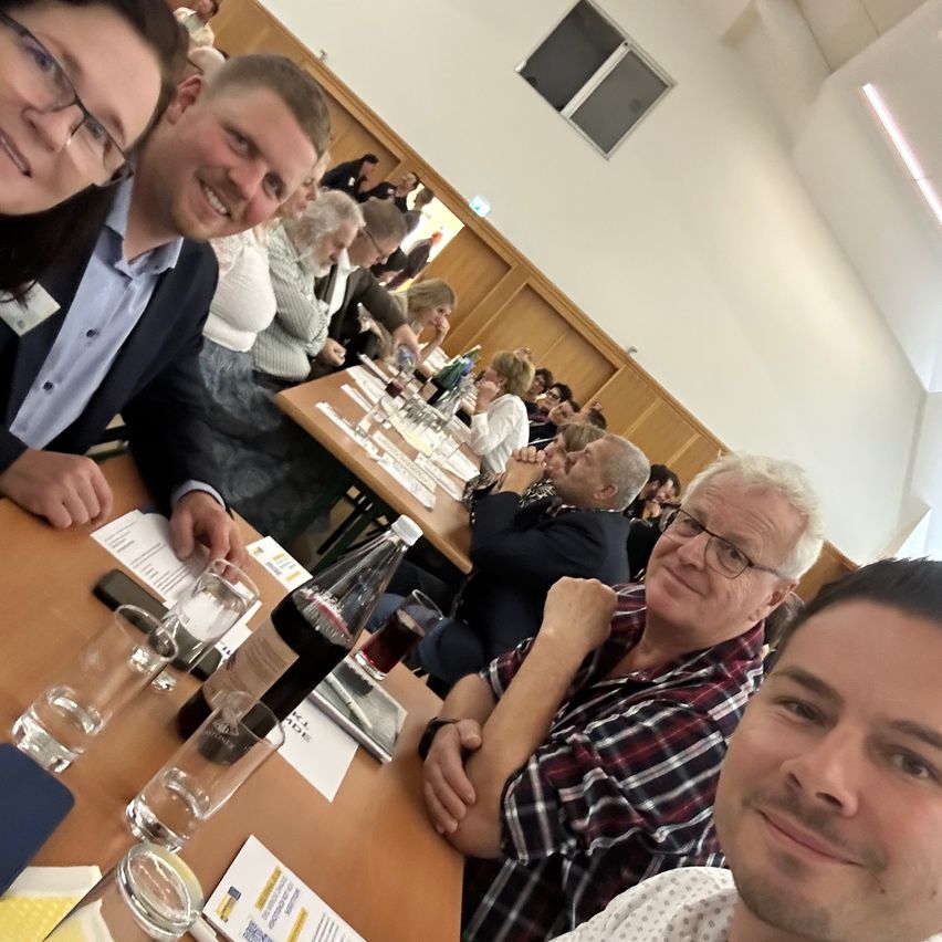 Mehrere Personen sitzen an Tischen in einem Raum, lächeln und posieren für ein Foto. Die Tische sind mit Gläsern, Flaschen und Papieren besetzt. Der Raum hat Holzwände und ein Fenster.