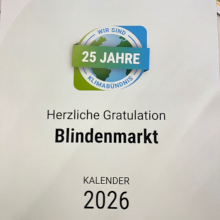 Ein weißer Plakat mit einem blau-grünen kreisförmigen Logo, das '25 Jahre Klimabündnis' besagt. Unten steht der Text 'Herzliche Gratulation Blindenmarkt'. Darunter steht das Wort 'Kalender', gefolgt von dem Jahr '2026'.