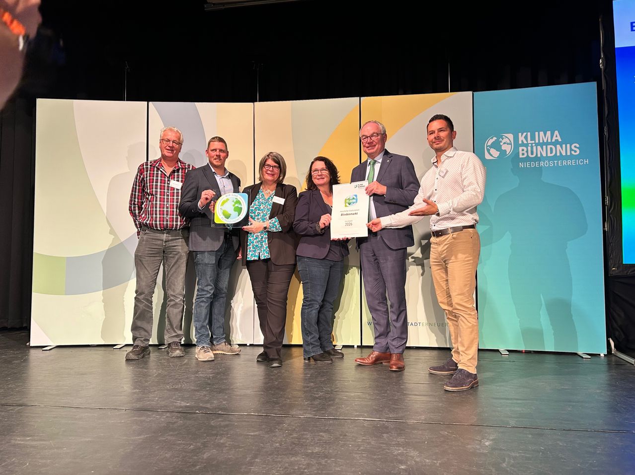 Fünf Personen stehen lächelnd auf einer Bühne und halten Urkunden in den Händen. Dahinter befinden sich Hintergrundkulissen mit Logos und Texten. Die Umgebung deutet auf eine Preisverleihung hin.