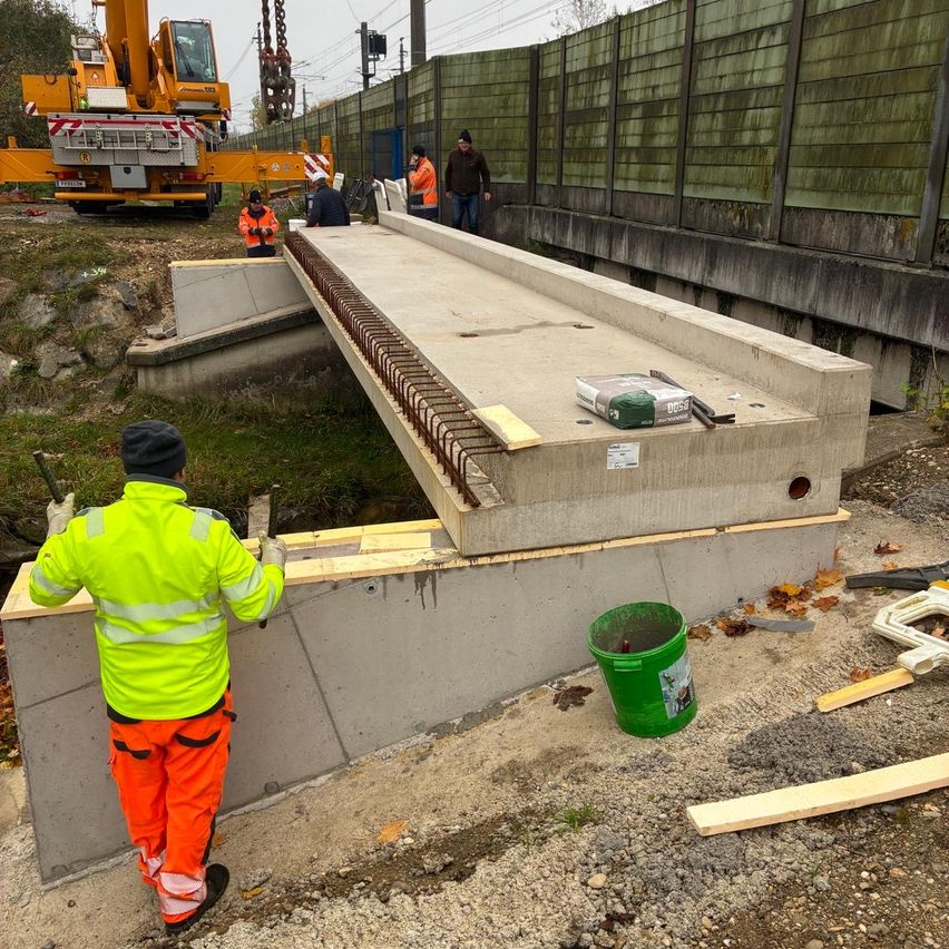 Bauarbeiter in leuchtender Schutzkleidung arbeiten an einer Betonbrücke unter einem Kran, mit einem grünen Eimer in der Nähe und einem Zaun dahinter.