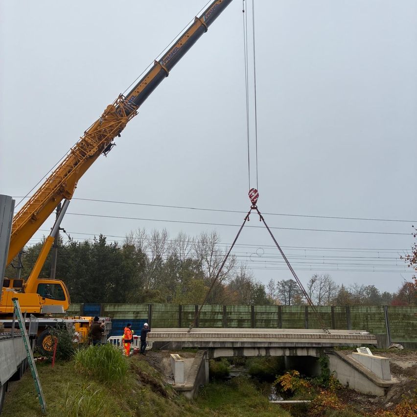 Ein großer Kran hebt eine Brücke an. Mehrere Arbeiter in orangefarbenen Westen und Helmen sind vor Ort. Der Kran ist gelb mit einem langen Ausleger. Die Szene spielt vor dem Hintergrund von Bäumen und Stromleitungen.