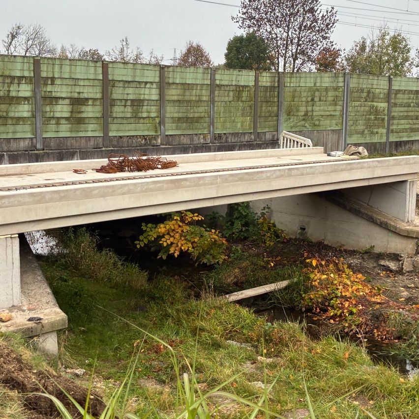 Eine Betonbrücke überspannt ein Gewässer mit verwilderten Pflanzen darunter. Eine rostige Kette liegt auf dem Brückendeck. Das Gebiet ist von einem grünen Metallzaun umschlossen.