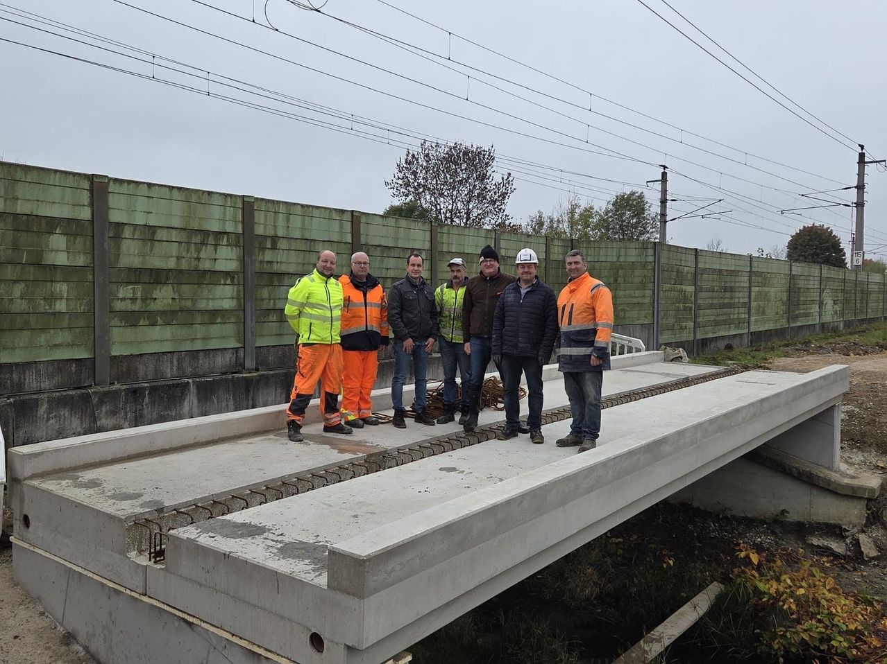 Eine Gruppe von Männern in Sicherheitskleidung steht auf einer Betonbrücke unter einem bewölkten Himmel. Hinter ihnen ist eine grüne Mauer und obere Drähte sichtbar.