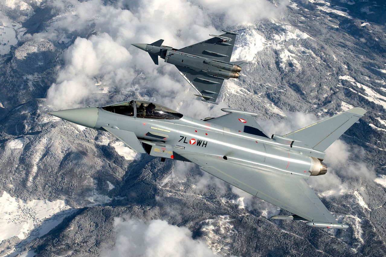 Zwei Militärjets fliegen im Himmel, einer hinter dem anderen, vor einer Kulisse aus verschneiten Bergen und Wolken. Der vordere Jet hat '7L WH' an seiner Seite.