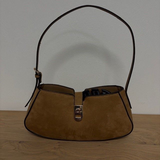 Eine braune Wildlederhandtasche mit schwarzem Henkel und silberner Schnalle steht auf einem Holztisch.