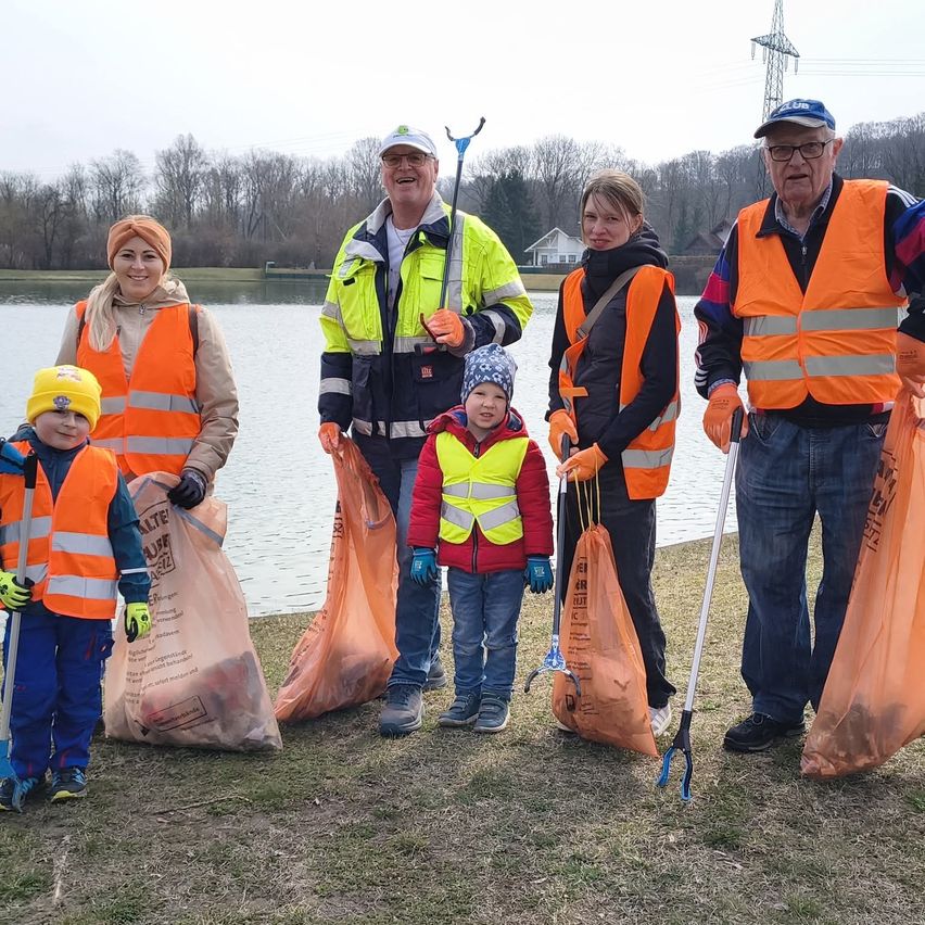 Eine Gruppe von Menschen in orangefarbenen Warnwesten, die Müllsäcke vor einem See halten. Sie lächeln und scheinen für ein Foto zu posieren.