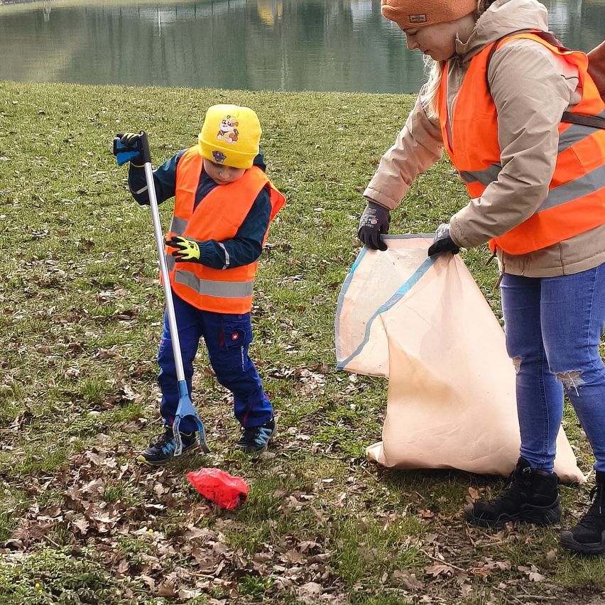 Ein junger Junge und eine Frau in Sicherheitswesten sammeln Müll am See. Der Junge hält eine Harke und die Frau eine große Tasche.