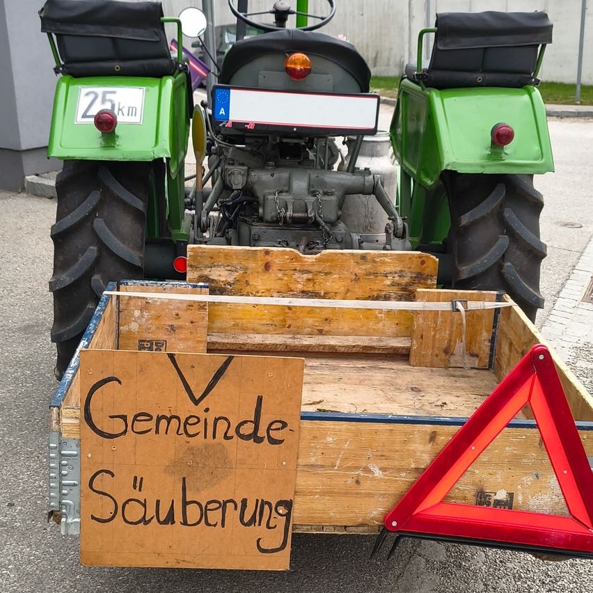 Ein grüner Traktor ist am Straßenrand geparkt, mit einem Holzkasten auf der Rückseite. Ein Schild auf dem Kasten steht Gemeinde Sauberung.