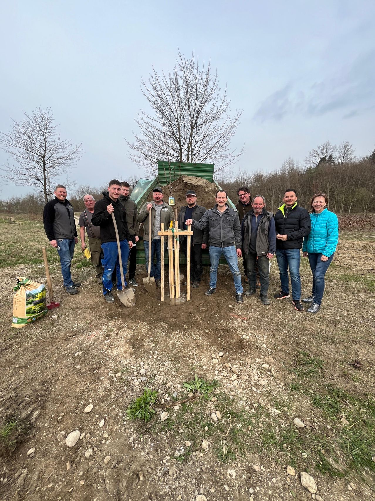 Eine Gruppe von Menschen steht um ein Loch herum und hält Schaufeln. Sie scheinen einen Baum zu pflanzen, mit einem grünen Container und Erde in der Nähe. Die Umgebung ist im Freien mit spärlicher Vegetation.