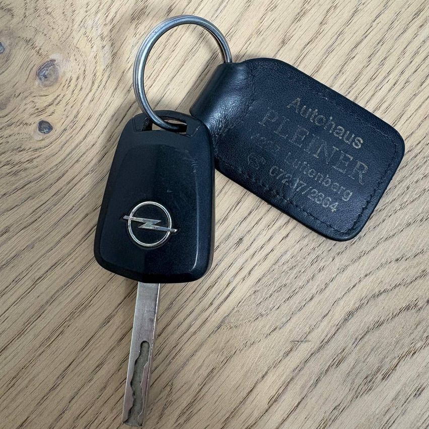 Ein schwarzer Opel-Schlüssel mit Schlüsselring liegt auf einer Holzoberfläche. Der Schlüsselring hat einen schwarzen Lederanhänger mit der Aufschrift 'Autohaus Pleiner' und Kontaktinformationen.