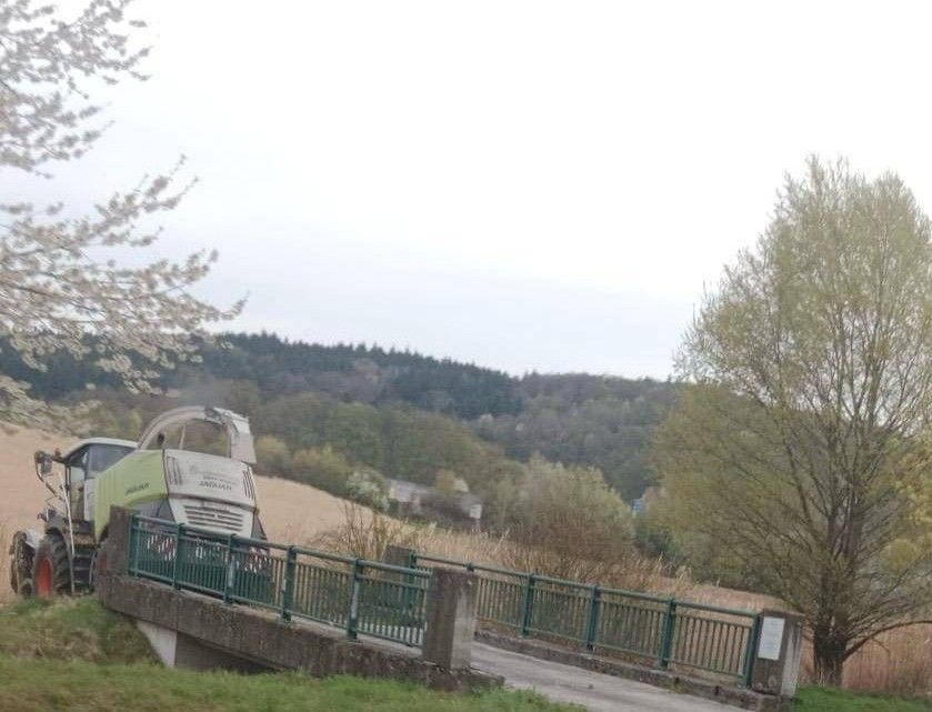Ein grüner und weißer Traktor steht auf einer Brücke mit grünen Geländern. Die Brücke befindet sich über einem Rasenbereich mit klarem Himmel darüber.