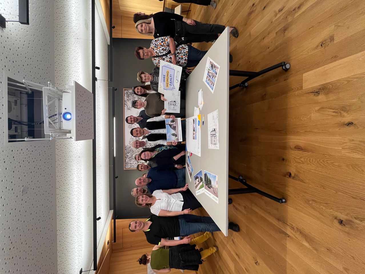 A group of people stand around a table in a room, holding certificates and smiling. Papers, photos, and a projector are on the table. The room has wooden floors and a projector on the left.
