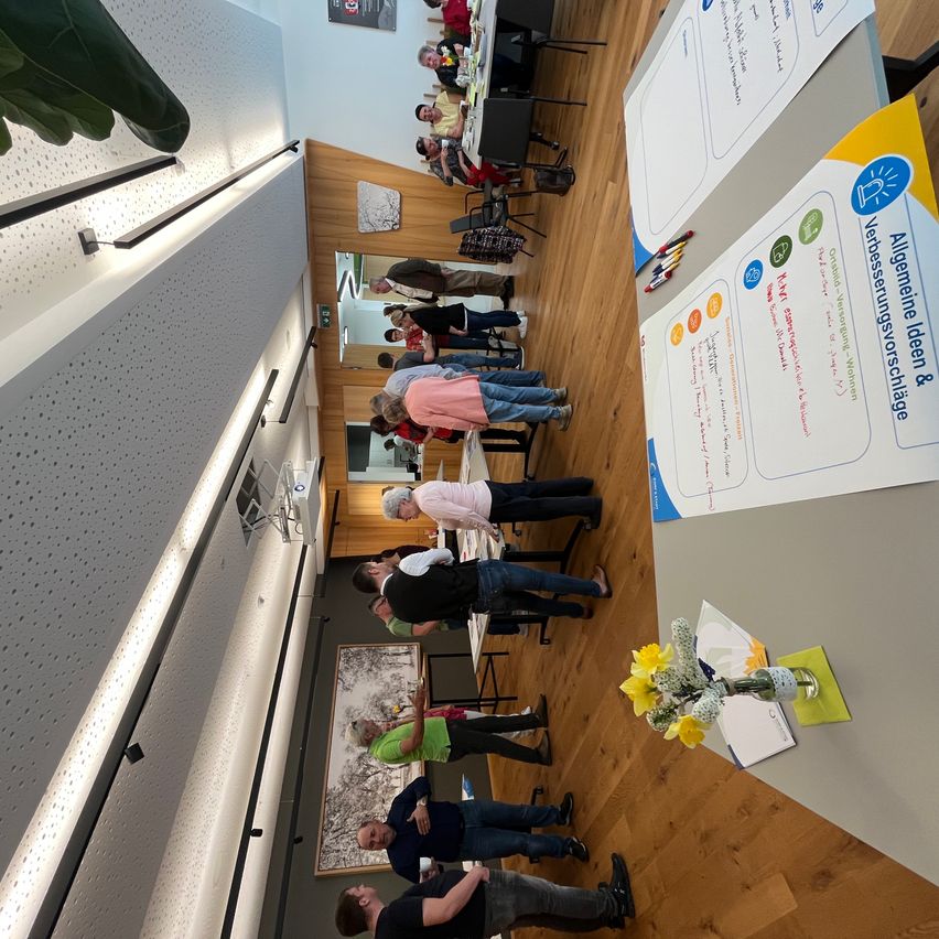 A group of people stand in a meeting room, engaging in discussions. Tables are set with papers and flowers. An overhead view shows the room's layout with wooden floors and white walls.