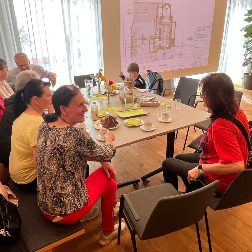 A group of people are sitting around a table with food, drinks, and a presentation on a projector screen. They are looking at the presentation.