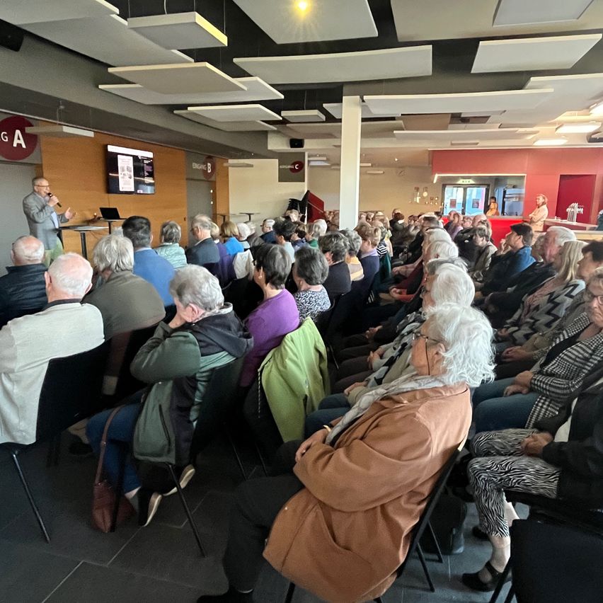 A speaker addresses a seated audience in a large room. The audience is predominantly elderly, and a projector screen is visible in the background.