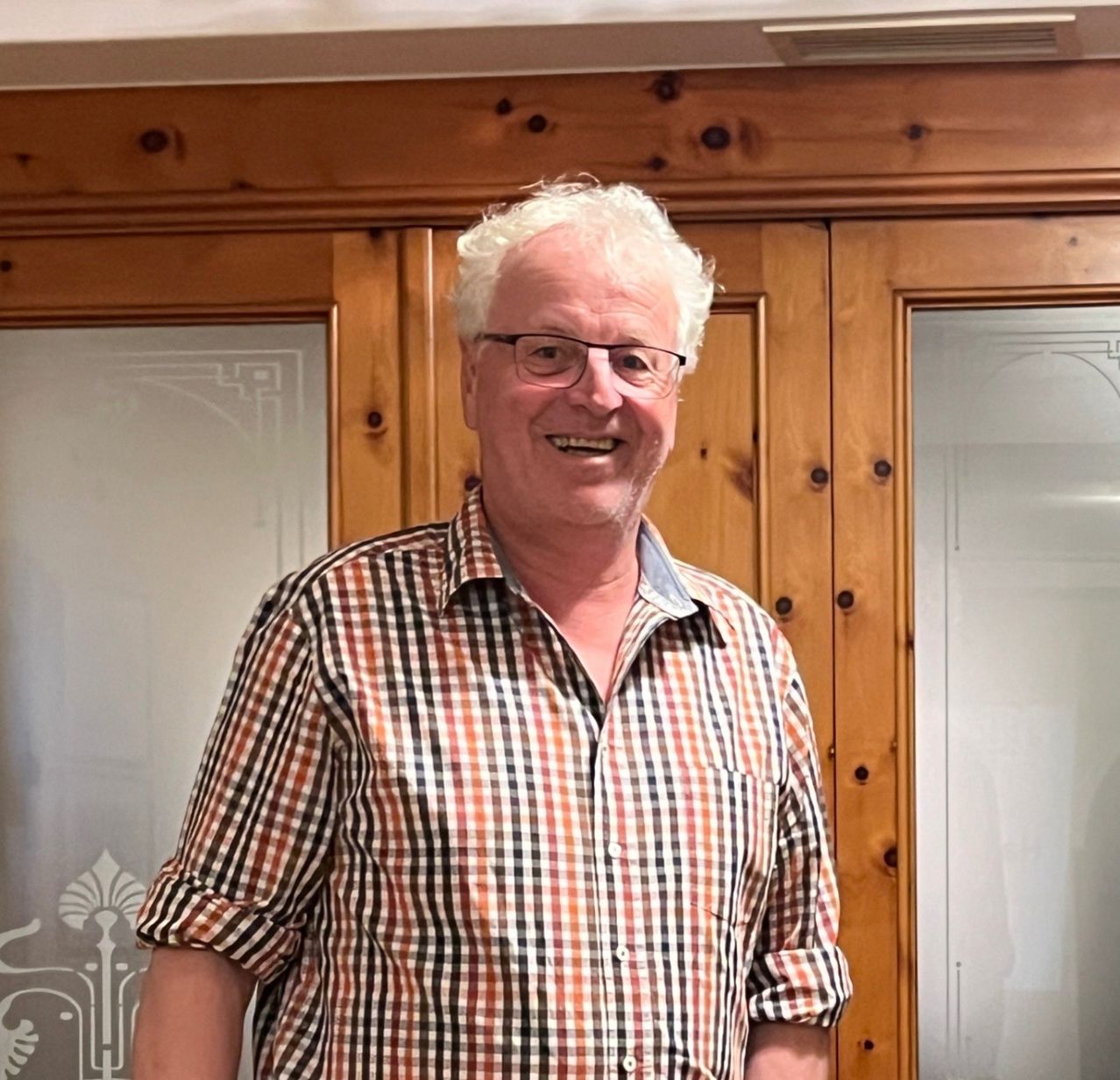 A man with white hair stands in front of wooden doors, smiling for the camera, wearing a checkered shirt.
