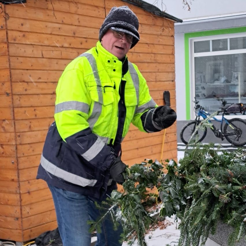 A man in a reflective yellow safety jacket stands in the snow, holding a branch of greenery, with a wooden building and parked bicycles in the background.