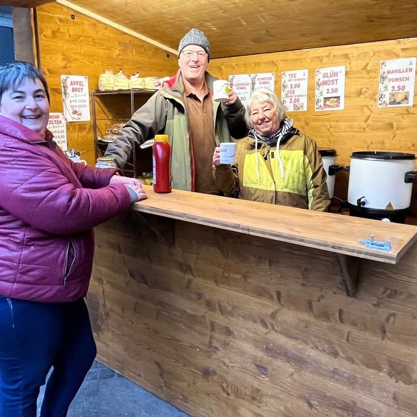 Three people are smiling and posing at a wooden counter. A man and woman hold cups, with a man in the middle. Behind them is a wooden wall with signs and a shelf.
