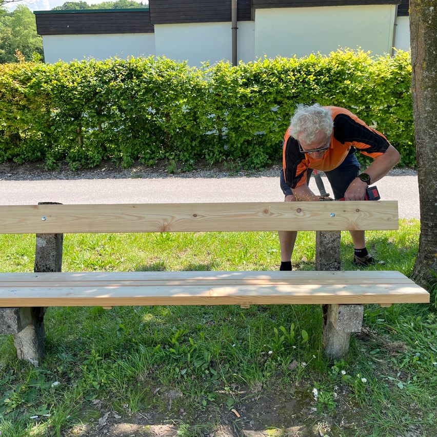 An elderly man in an orange shirt works on a wooden bench with a power drill. The bench is set on concrete in a grassy area.