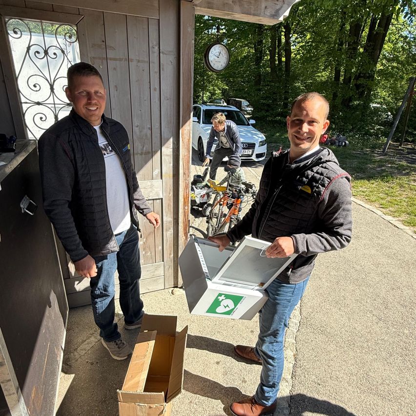 Two men are standing outside a wooden door, one holding a box. A bicycle and a car are in the background. Another man is working on the bicycle.