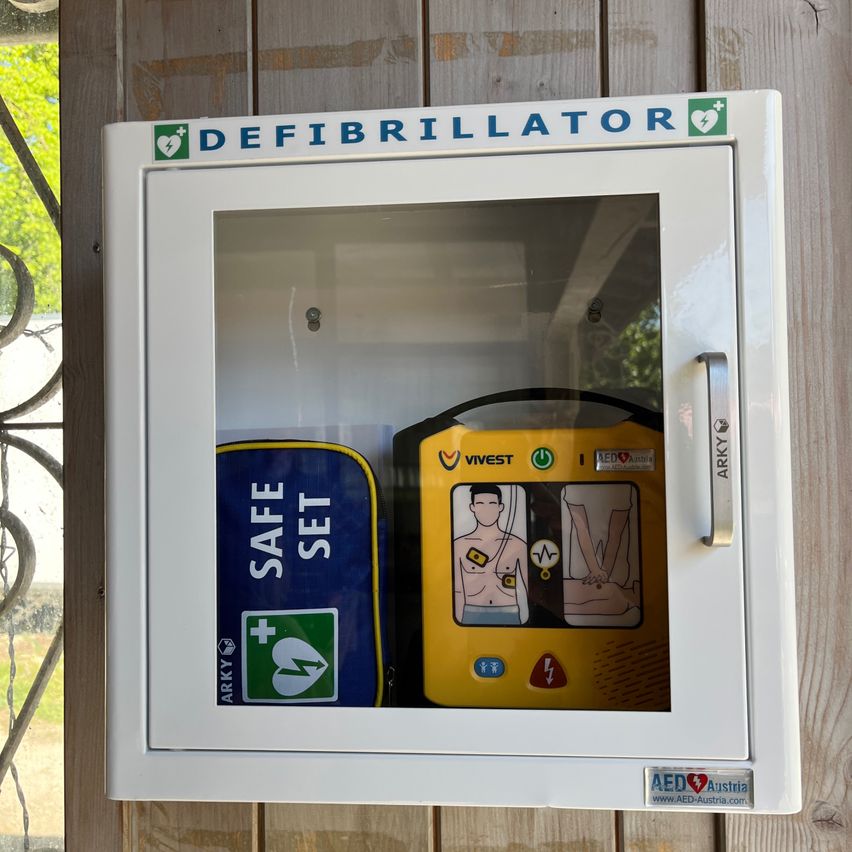A white defibrillator cabinet with a glass door, labeled 'Defibrillator'. Inside is a yellow device with a safety set in a blue case.