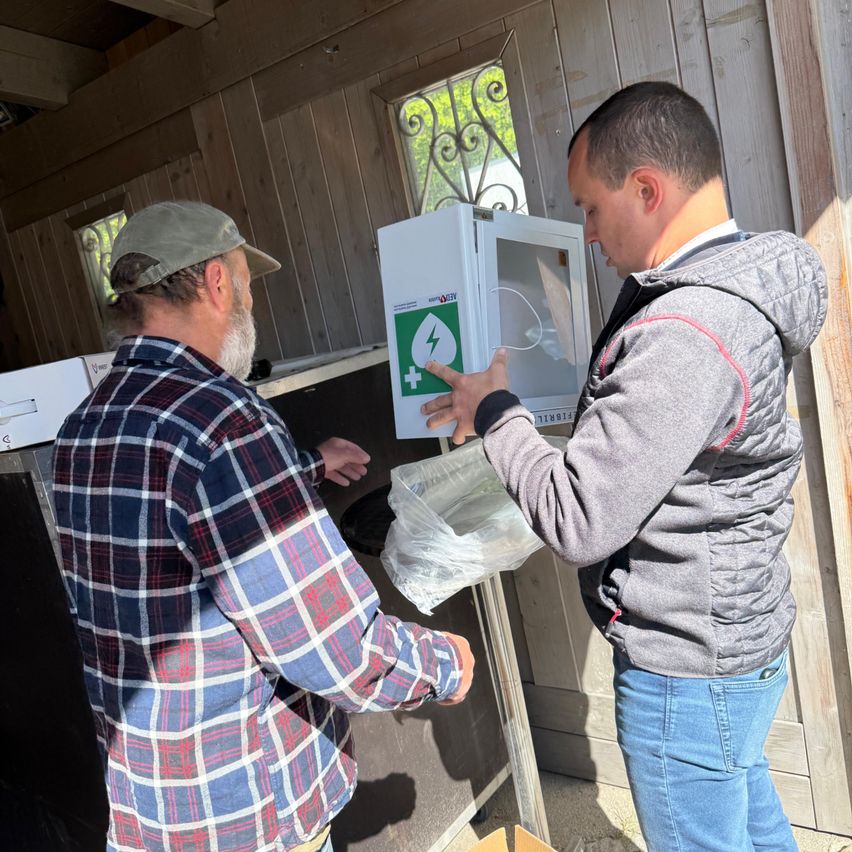 Two men are in a shed, one holding a white box with a green symbol, and the other holding a plastic bag. The box has an open door.