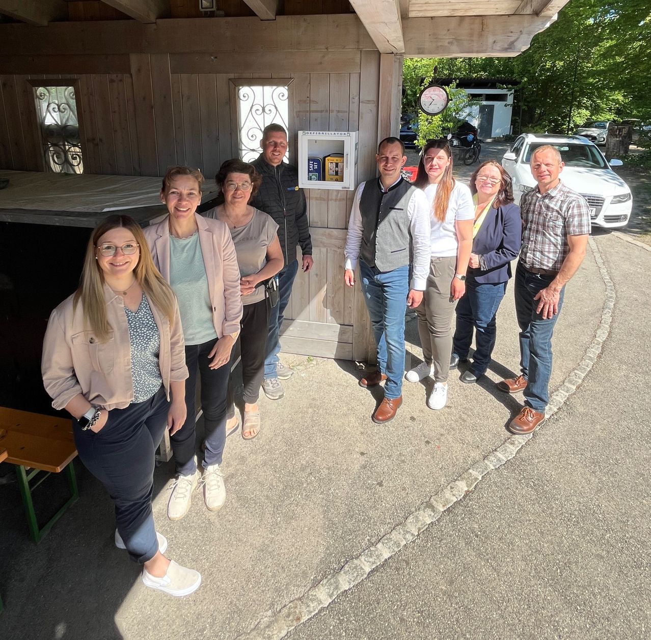A group of people are posing for a photo outside a wooden shed with a sign and a car parked nearby.