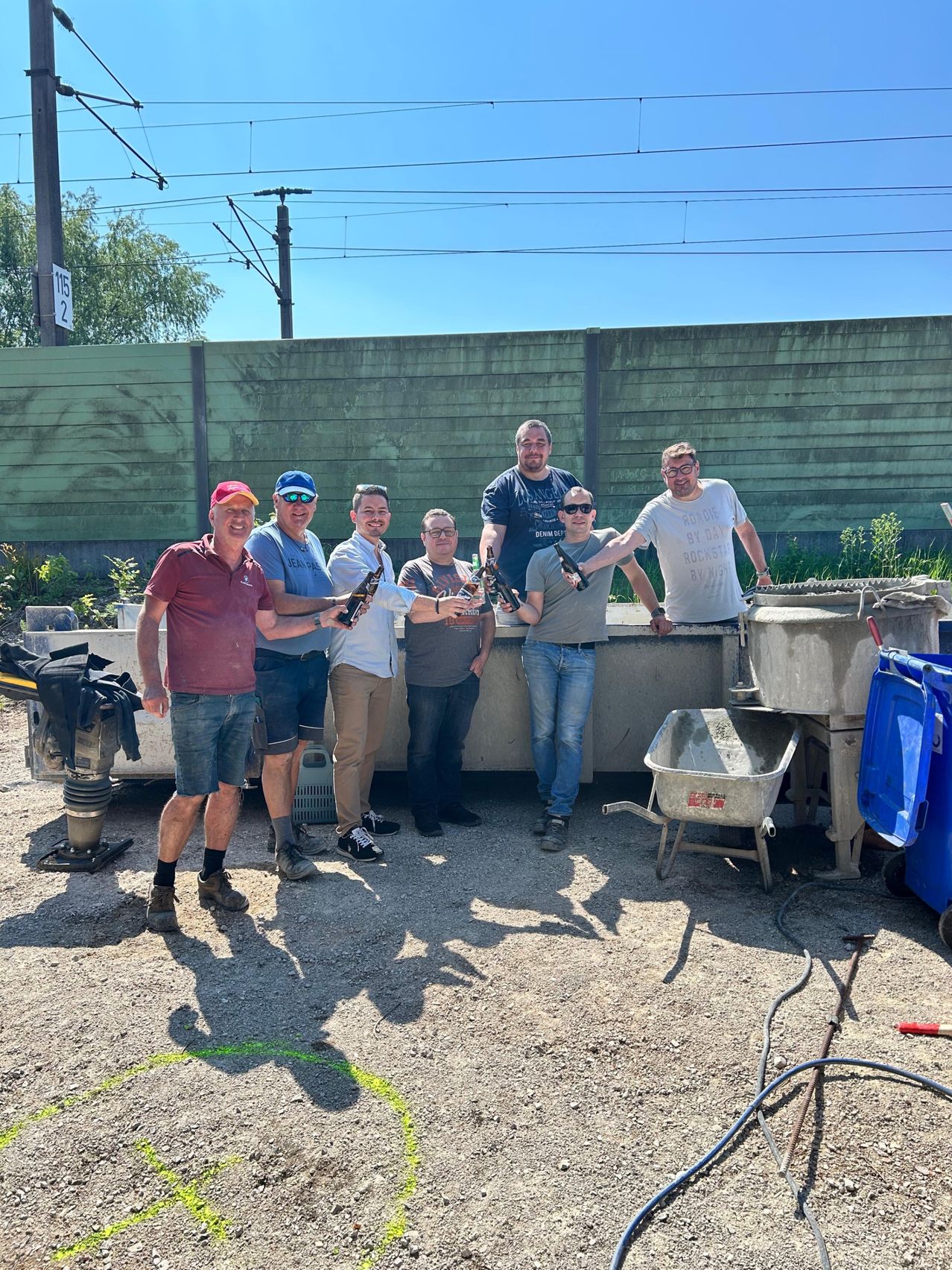 A group of men stand together holding bottles, posing for a photo in front of a construction site with a wall and some equipment.
