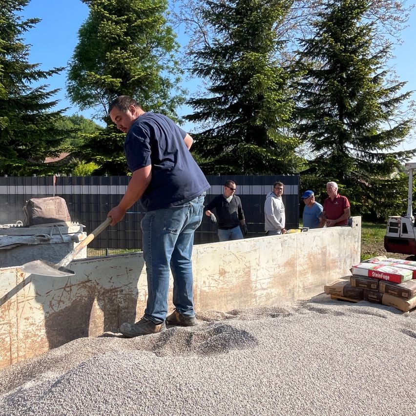 A man is shoveling gravel in an outdoor construction site with others watching. A wall, a pile of bricks, and a fence are in the background.