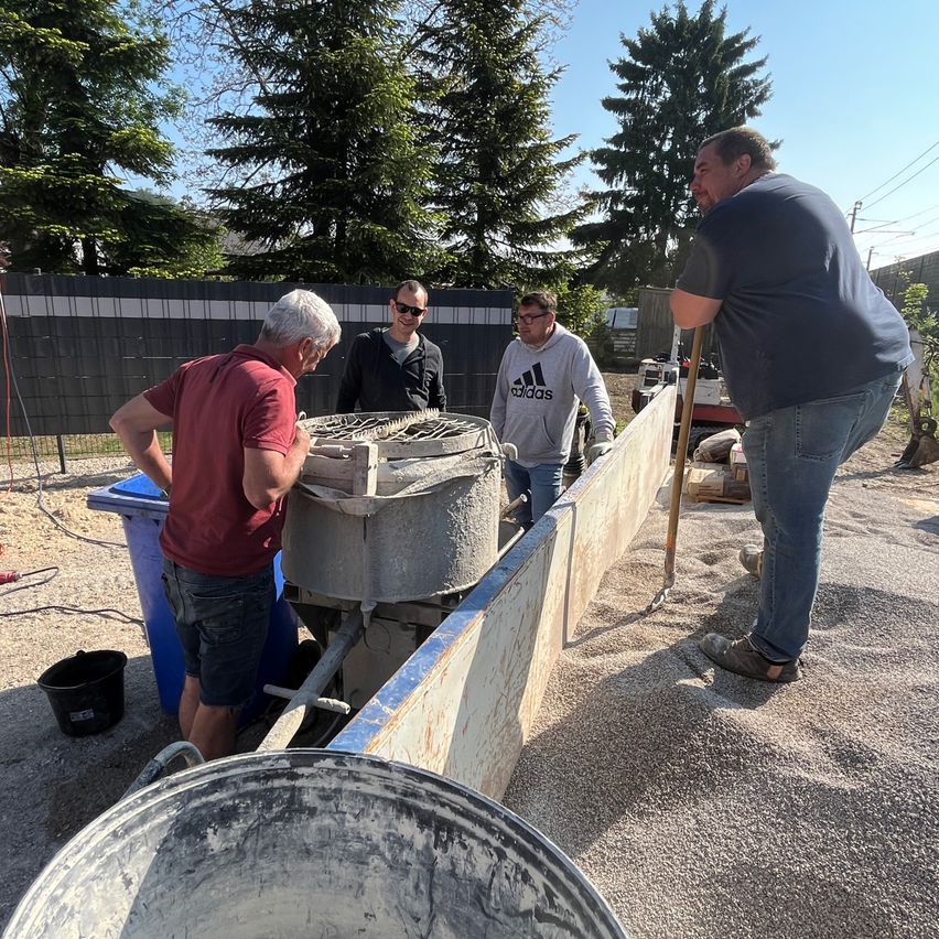 Four men in casual clothes are working on a concrete wall with buckets and tools. Trees and a fence are in the background.