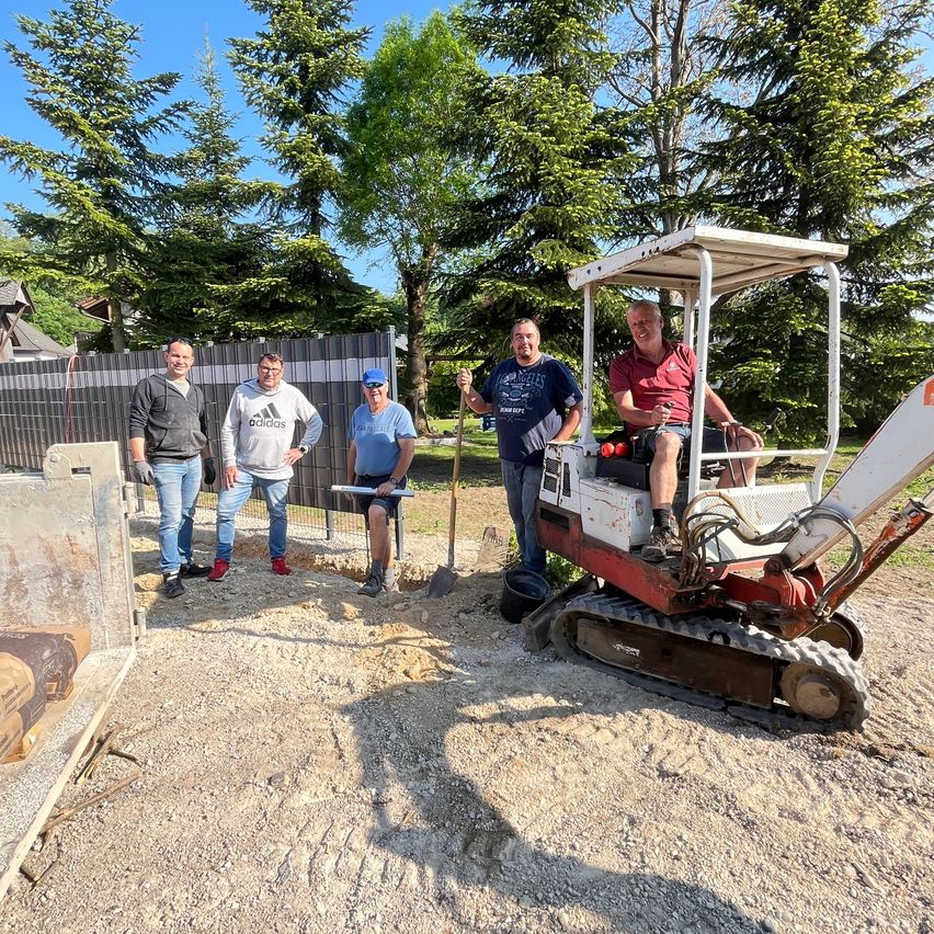 Five men are standing on a construction site. One is sitting on a backhoe. One man holds a shovel, another holds a rake.