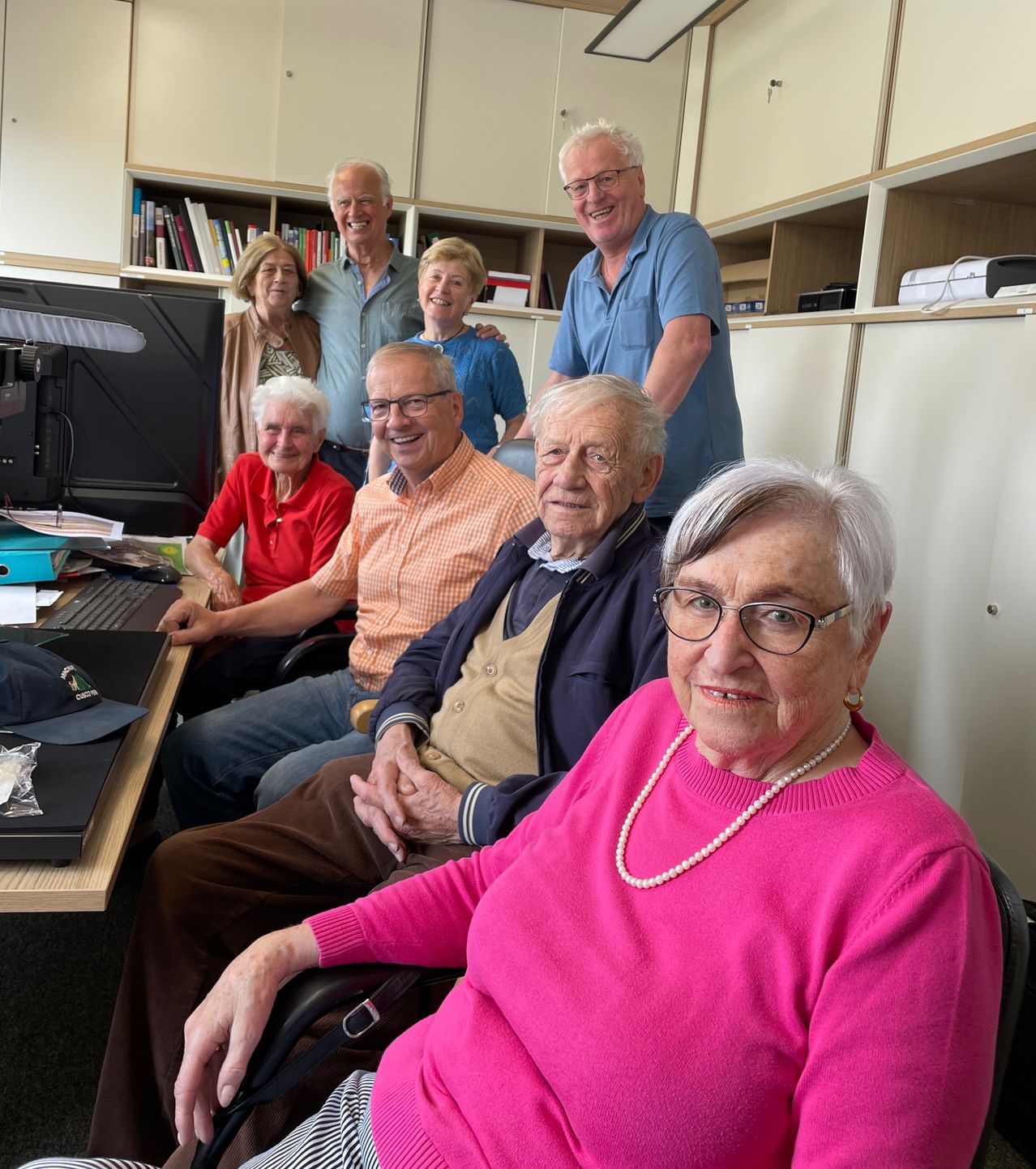 Eine Gruppe älterer Menschen lächelt und posiert für ein Foto in einem Büro. Vor ihnen steht ein Schreibtisch mit einem Computer.