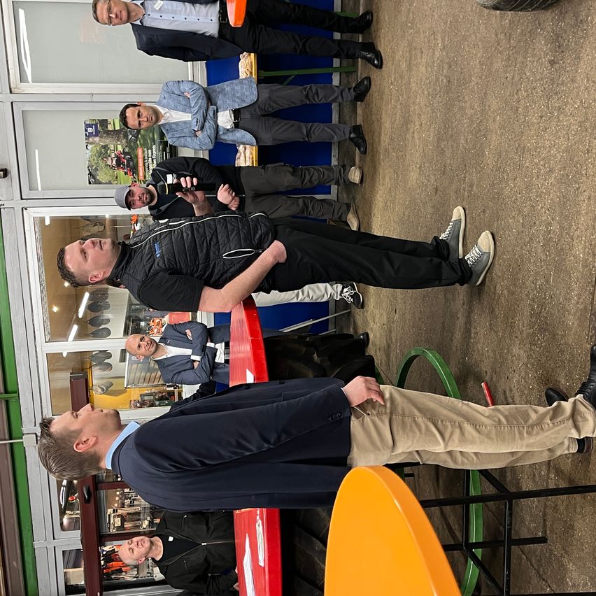 Mehrere Männer stehen in einem Innenbereich. Ein Mann hält ein Mikrofon. Ein anderer Mann lehnt sich an einen Tisch. Der Raum hat eine Glaswand und einige Gegenstände sind ausgestellt.