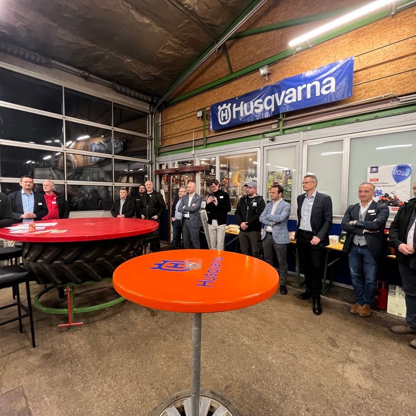 Eine Gruppe von Menschen steht in einem Raum mit einem Husqvarna-Banner an der Wand. Sie tragen formelle Kleidung. Es gibt zwei Tische mit Gegenständen darauf. Ein Tisch hat einen Reifen darauf. Es gibt Lampen an der Decke.