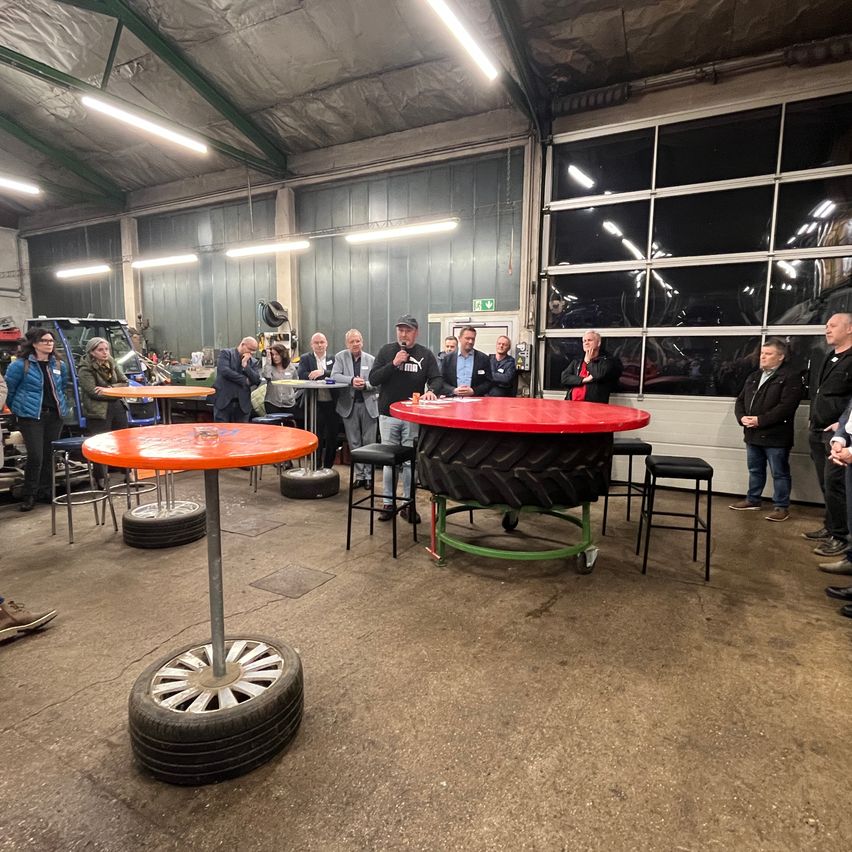 Eine Gruppe von Menschen versammelt sich in einer großen Garage, einige stehen, andere sitzen um Tische herum. Reifen werden als Tischgestelle wiederverwendet. Ein Mann spricht in ein Mikrofon am zentralen Tisch.