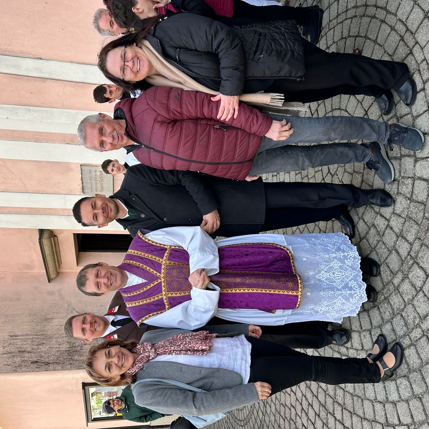 Eine Gruppe von Menschen steht zusammen auf einer Kopfsteinpflasterstraße und lächelt für ein Foto. Der Mann in der Mitte trägt eine purpurne und goldene Weste, während andere Winterkleidung tragen.