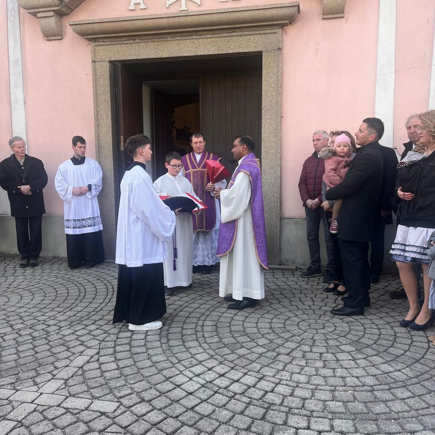 Vor einer Kirche findet eine religiöse Zeremonie statt. Ein Priester und zwei Messdiener stehen und halten ein Buch. Menschen stehen um sie herum.