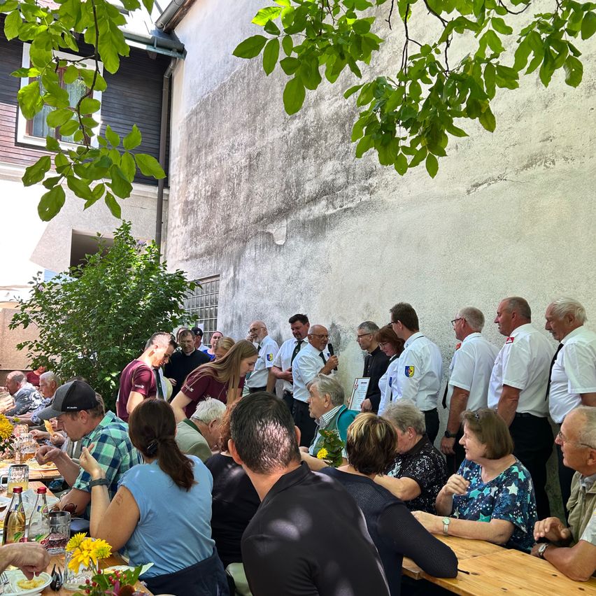 Eine Gruppe von Menschen, einige in Uniformen, versammelt sich um Tische in einer Außenumgebung, möglicherweise für eine Gemeinschaftsveranstaltung. Bäume und ein Gebäude sind im Hintergrund sichtbar.