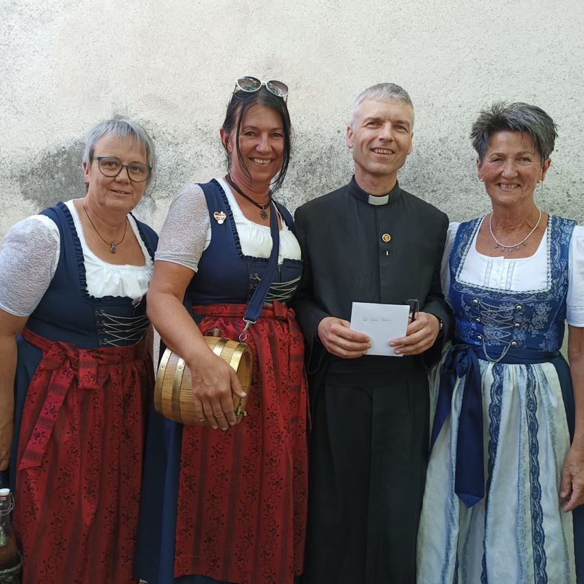 Vier Menschen lächeln und posieren für ein Foto. Drei Frauen tragen traditionelle deutsche Kleider, und ein Mann ist in einem schwarzen Klerikergewand gekleidet.