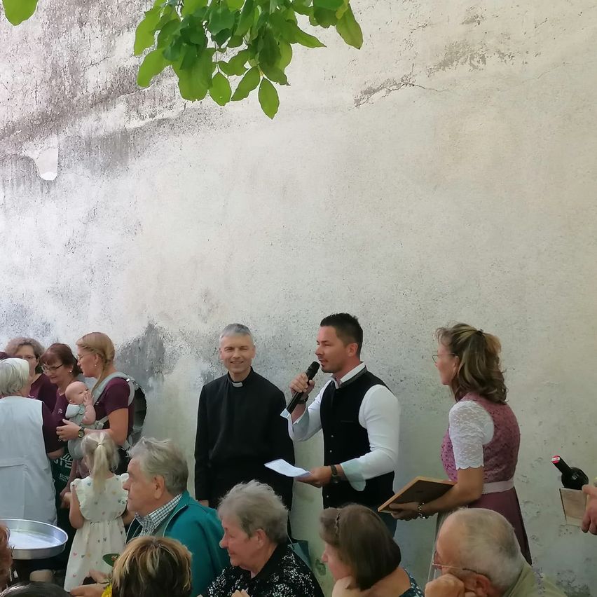 Eine Gruppe von Menschen versammelt sich im Freien, möglicherweise für eine Zeremonie. Ein Mann hält ein Mikrofon und spricht, während andere aufmerksam zuhören. Eine Frau hält ein Baby und ein Mann in einem schwarzen Gewand steht in der Nähe.