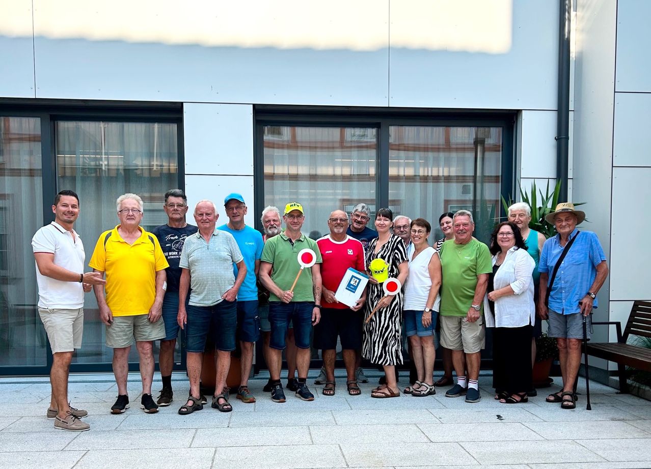 Eine Gruppe von Menschen posiert für ein Foto vor einem Gebäude mit großen Fenstern. Sie alle lächeln und einige halten rote Flaggen.