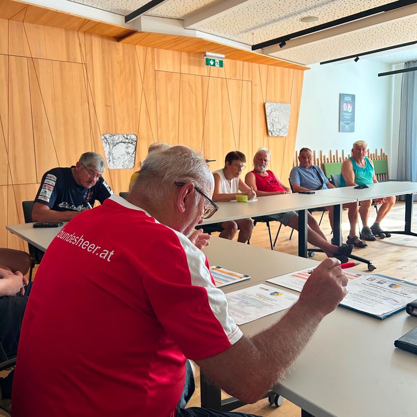 Eine Gruppe älterer Männer sitzt in einem Konferenzraum an einem langen Tisch und trägt Freizeitkleidung. Ein Mann in einem roten Shirt schreibt auf ein Stück Papier.