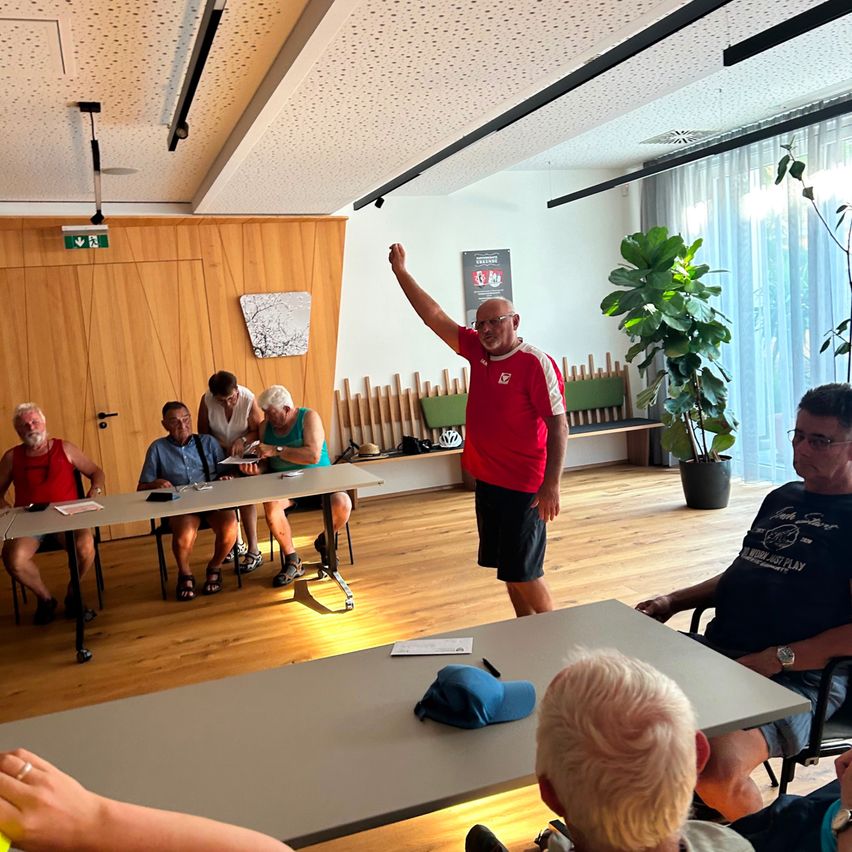 Eine Gruppe älterer Männer sitzt an einem Tisch in einem Raum. Ein Mann in einem roten Shirt steht vor ihnen. Papiere und eine blaue Mütze liegen auf dem Tisch. Eine Topfpflanze steht in der Ecke.