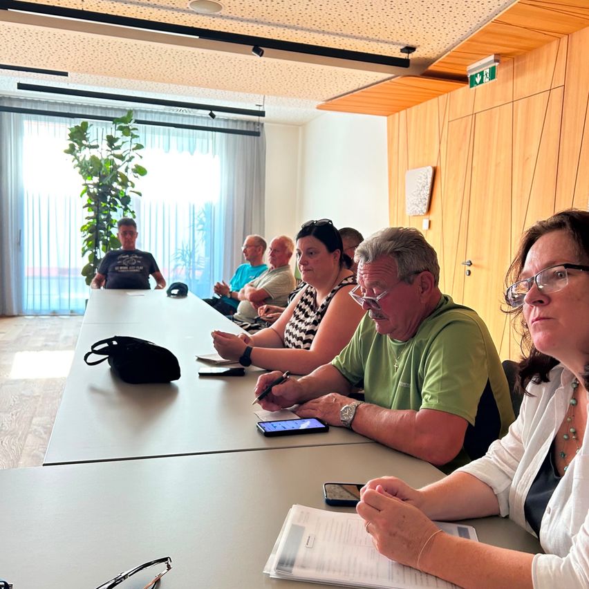 Eine Gruppe von Menschen sitzt in einem Besprechungsraum um einen Tisch herum, einige notieren etwas, während andere auf ihre Handys schauen. Eine Frau mit Brille hält einen Stift und Papier.